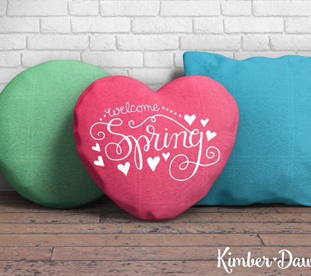 A round green pillow, a blue square pillow and a red heart shaped pillow that says, "Welcome Spring" all sitting on a wooden bench up against a white brick wall