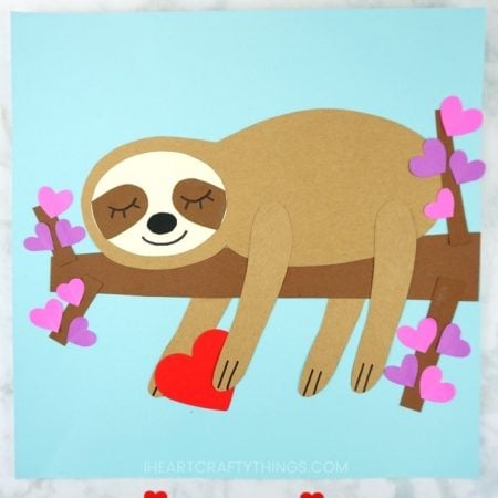 A sloth laying on a tree branch holding a red heart