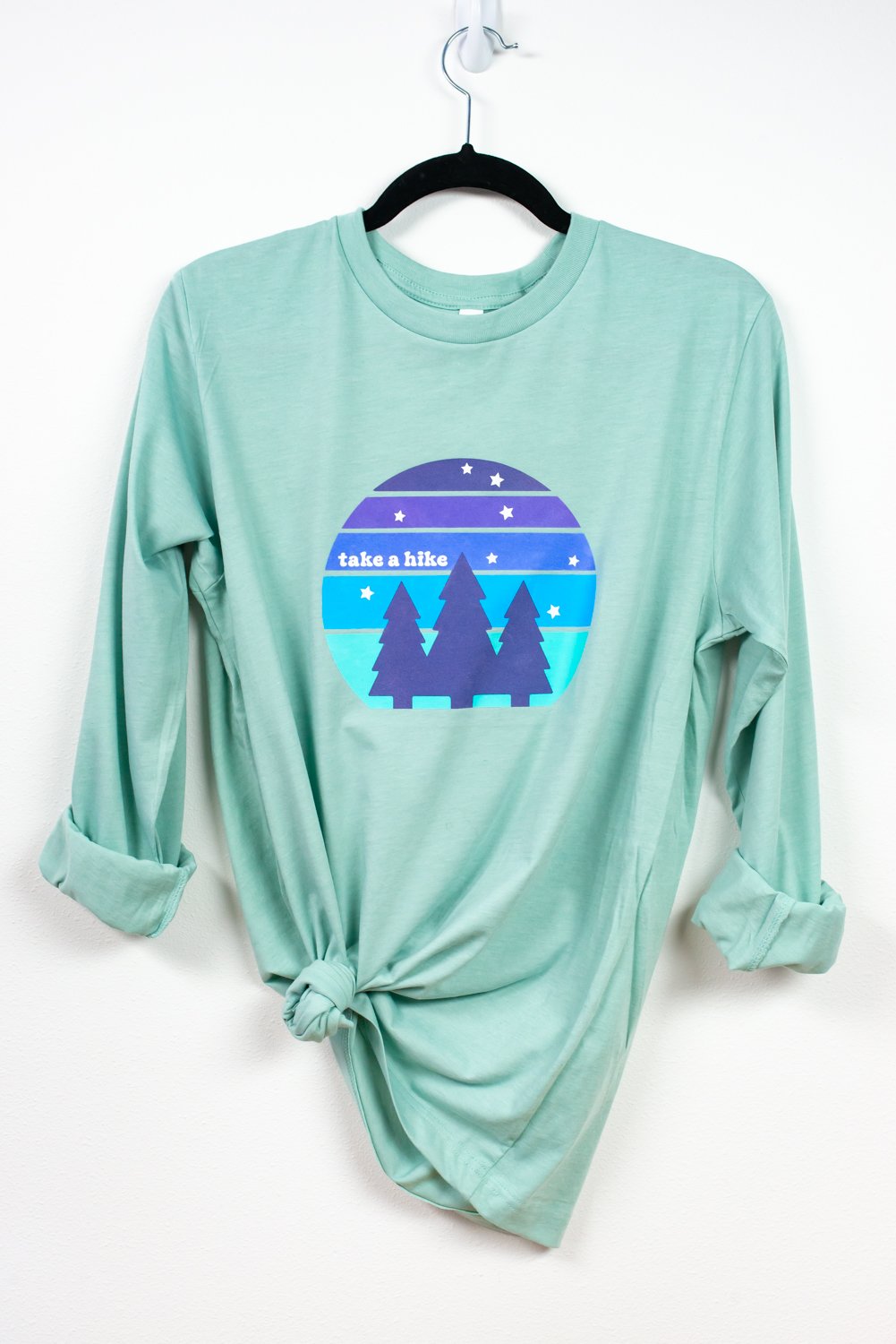 Mint-colored shirt with "take a hike" image hanging from hanger on white wall.