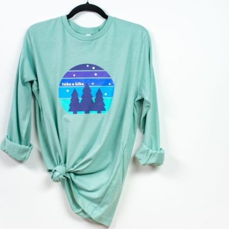Mint-colored shirt with "take a hike" image hanging from hanger on white wall.