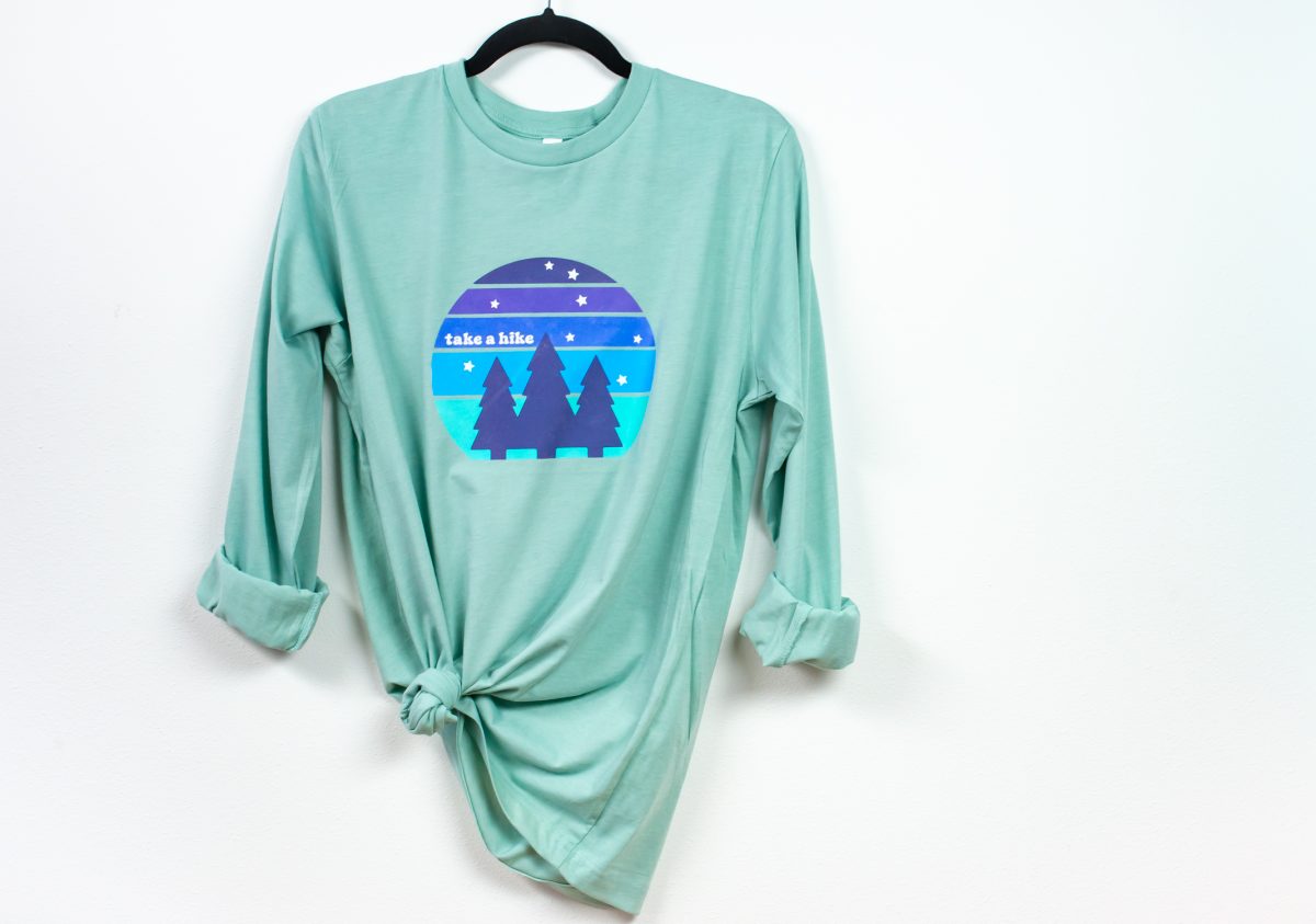 Mint-colored shirt with "take a hike" image hanging from hanger on white wall.