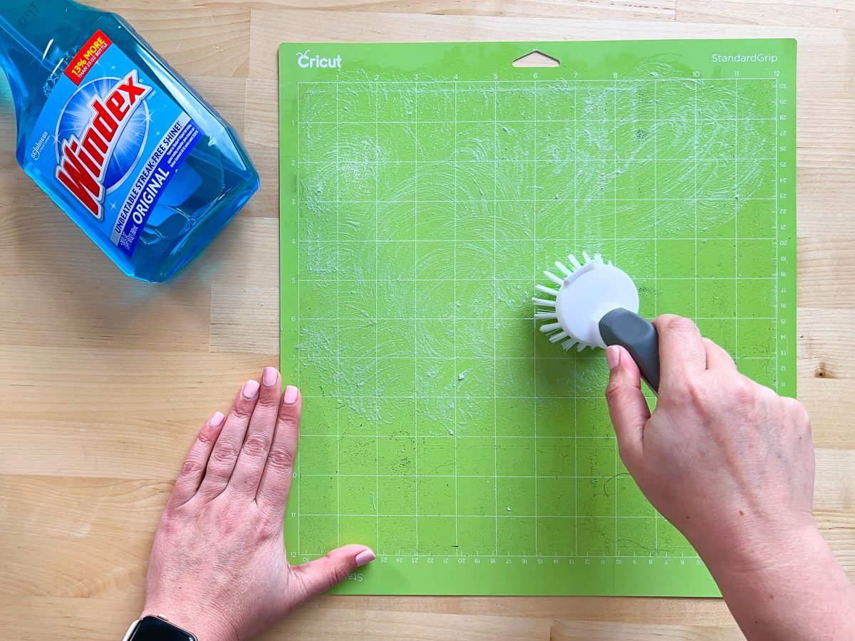 Overhead shot of hands using a bristled brush and Windex to clean a Green cricut mat.