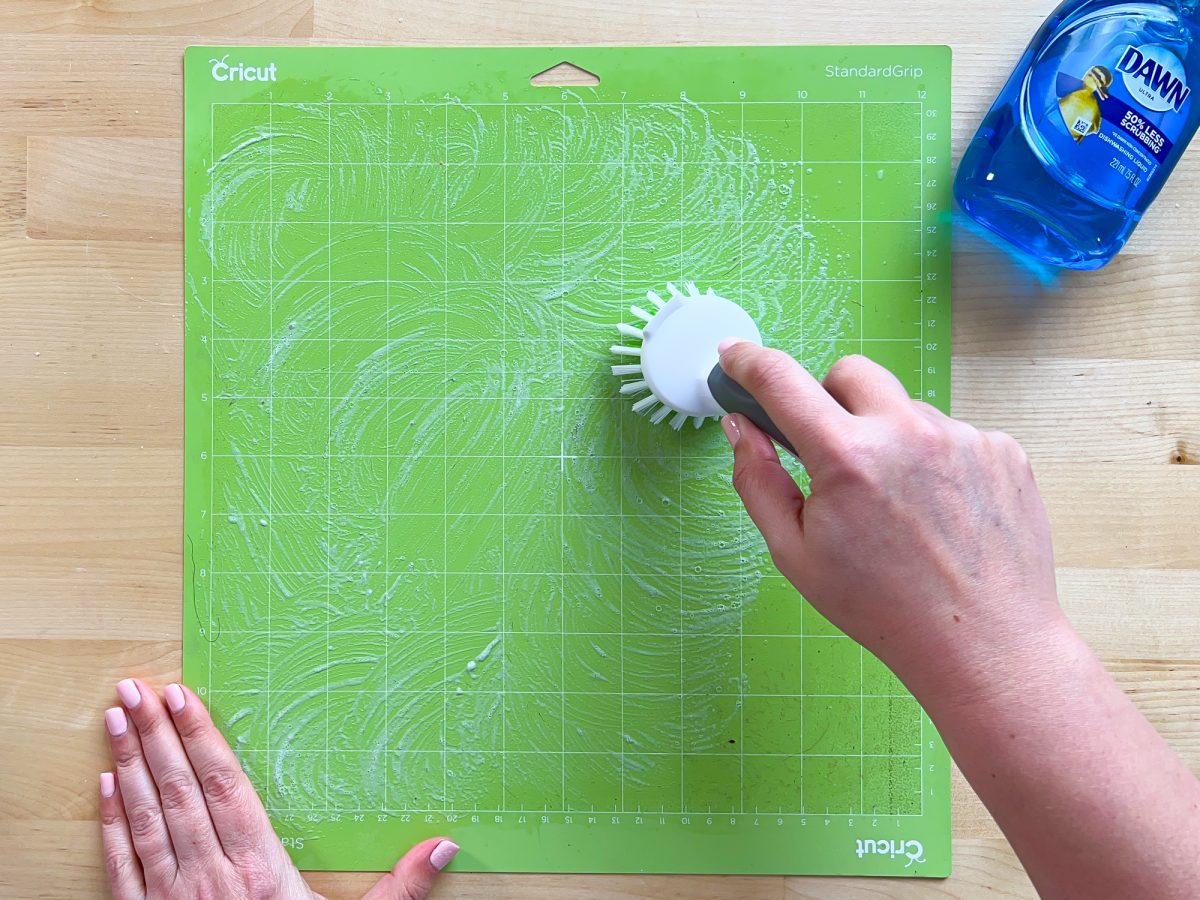 Overhead shot of hands using a bristled brush and Dawn to clean a Green cricut mat.