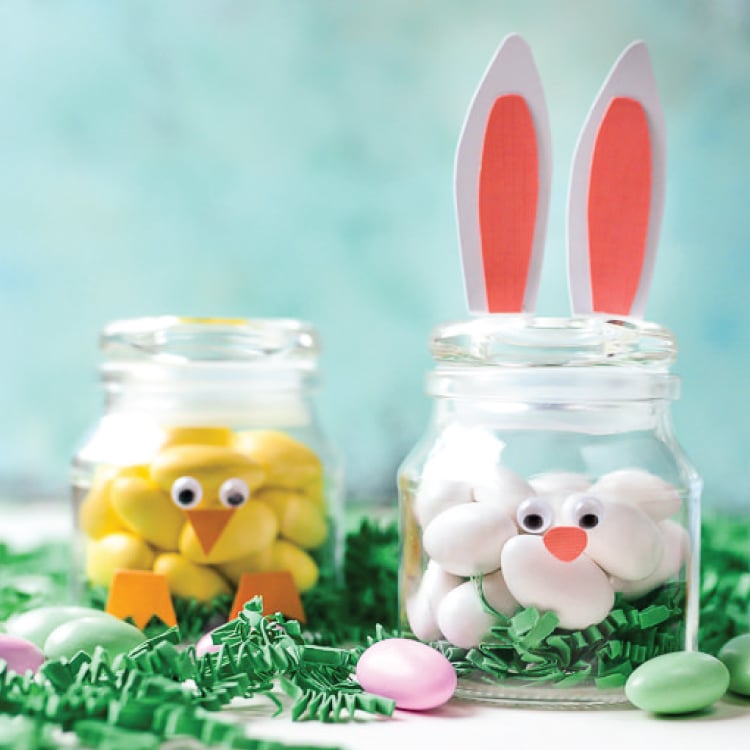 Three small jars on a table surrounded by greenery and Easter candy.  The jars are decorated to resemble a baby chick, a rabbit and a bird