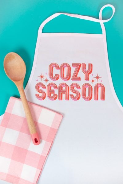 Sublimation apron with "cozy season" image. Pink checkered napkin, wooden spoon. Teal background