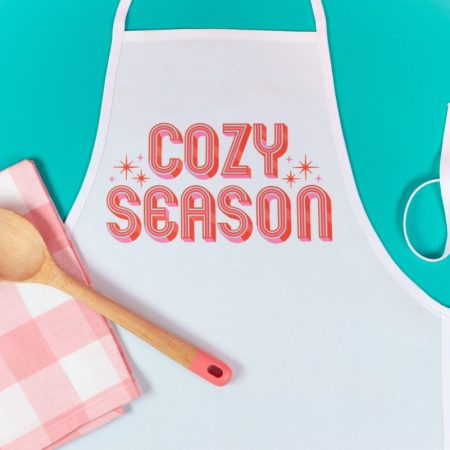 Sublimation apron with "cozy season" image. Pink checkered napkin, wooden spoon. Teal background