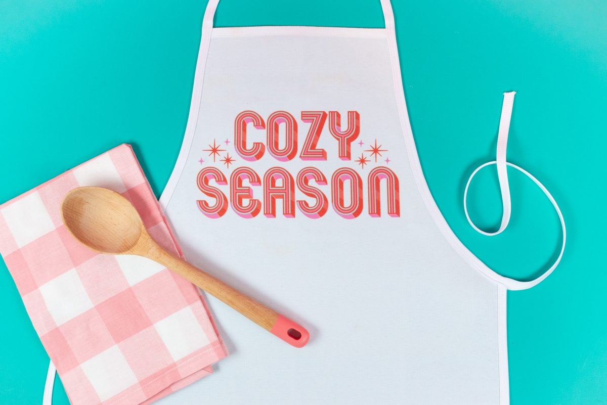 Sublimation apron with "cozy season" image. Pink checkered napkin, wooden spoon. Teal background