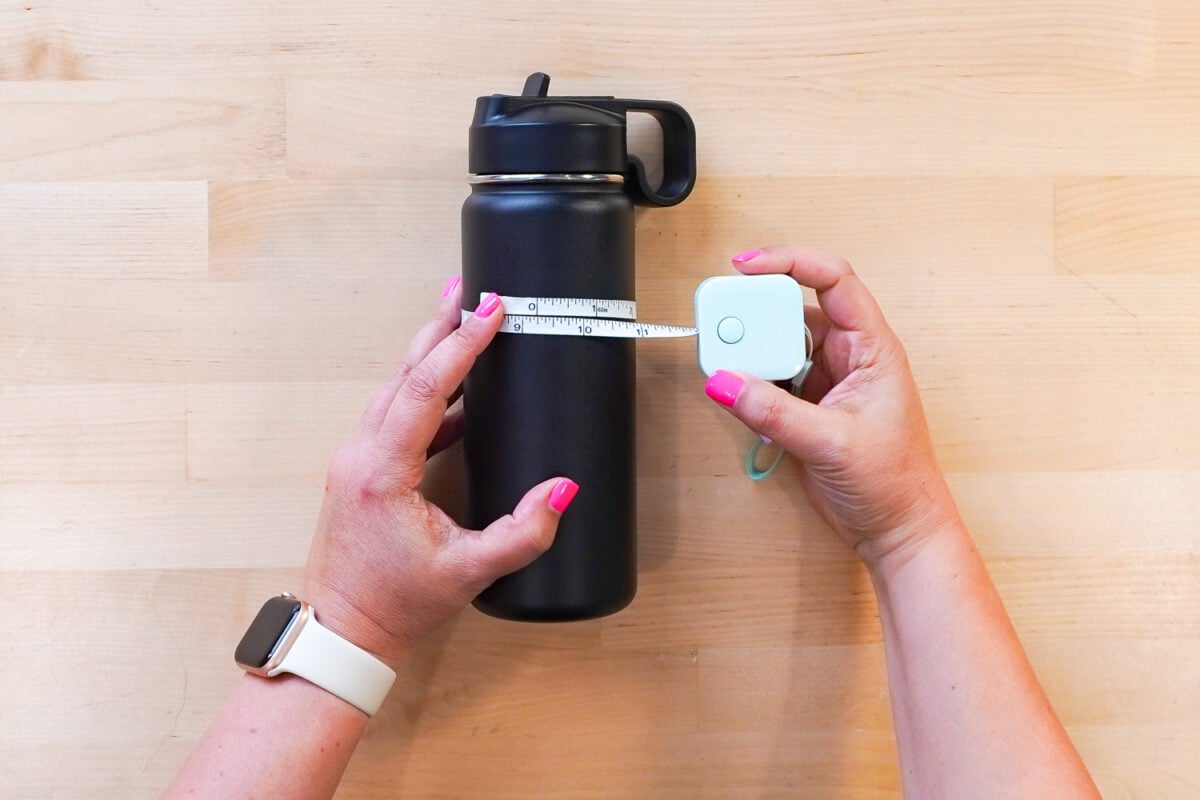 Hands measuring the circumference of a black tumbler using a tape measure