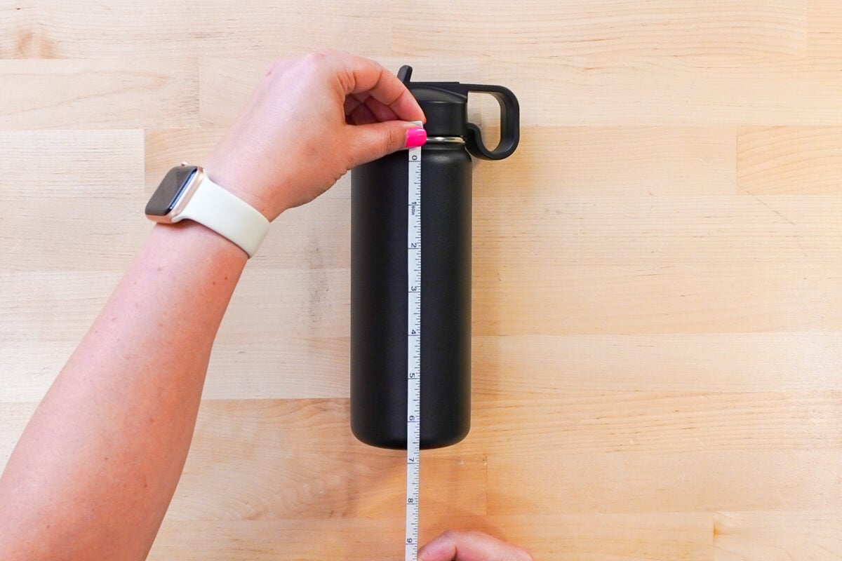 Hands measuring the height of a black tumbler using a tape measure