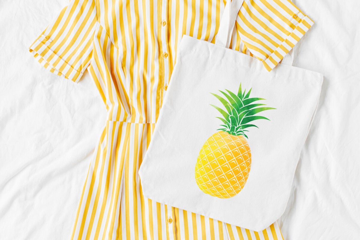 white tote bag with pineapple image on it, styled with a yellow striped dress.
