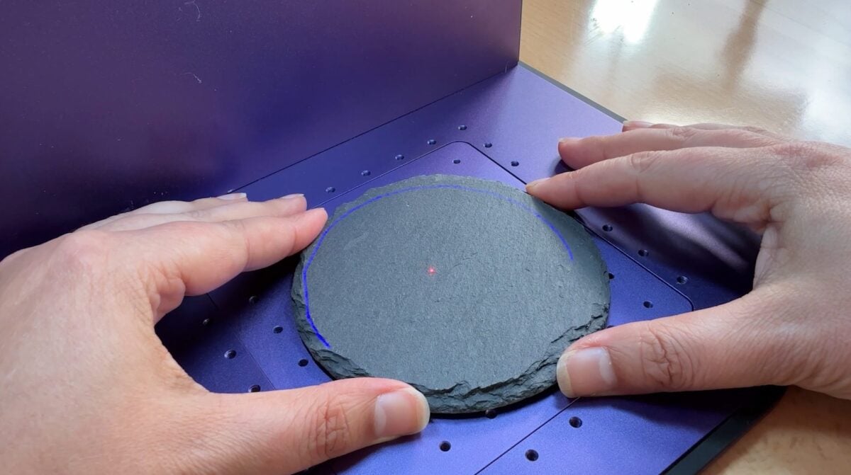 Slate coaster in laser, with hands moving the coaster into the blue framing circle.