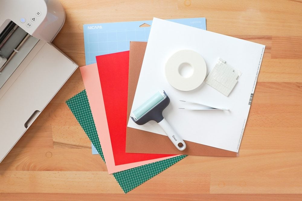 Table with supplies: Cricut Maker 4, blue mat, five colors of cardstock, foam adhesive, brayer, and tweezers